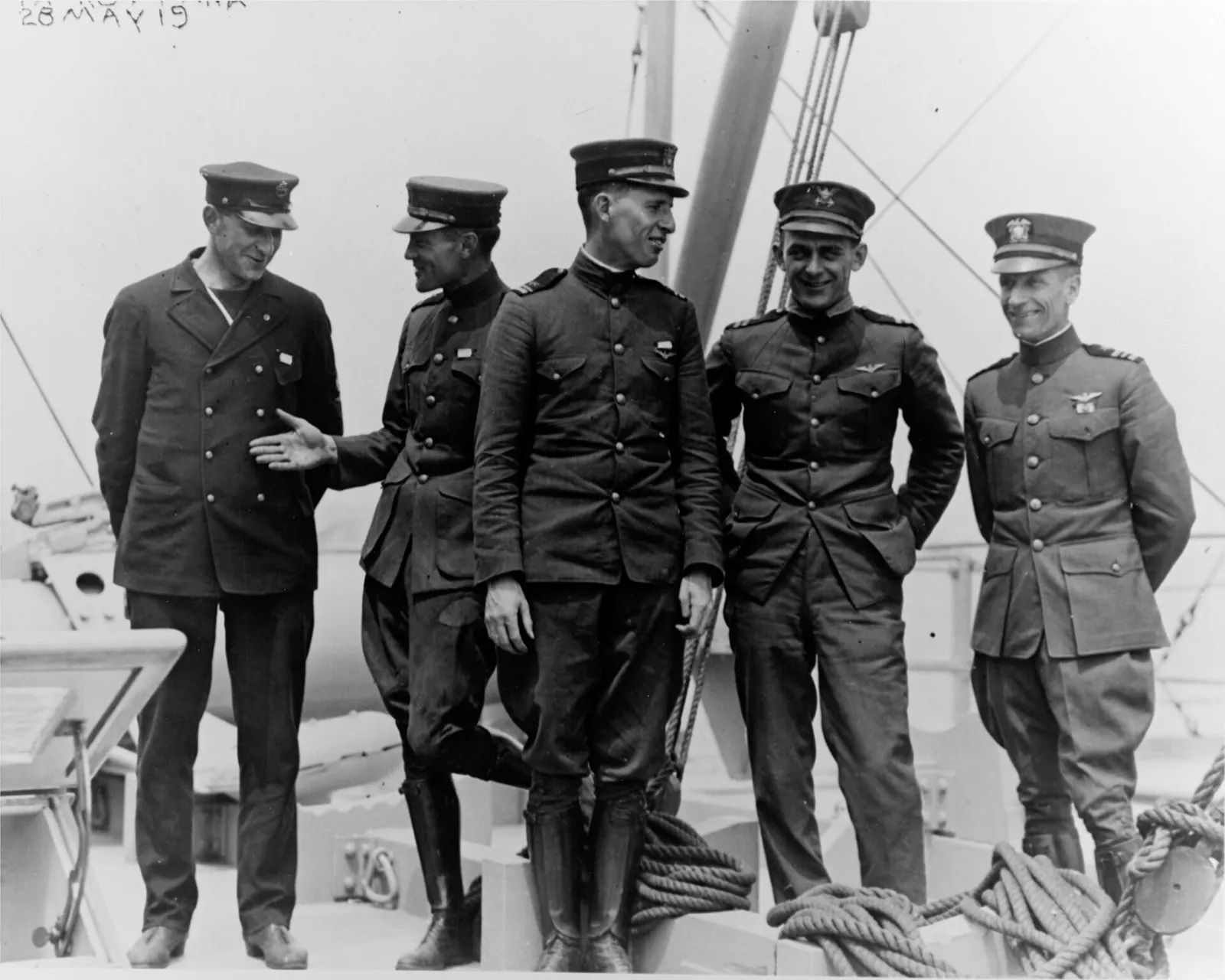 Five men in naval uniforms and peaked caps stand in a row on the deck of a ship, by a coil of rope and rigging. The man on the left wears a dark double-breasted jacket; the others wear high-collared tunics, and two of them riding boots.