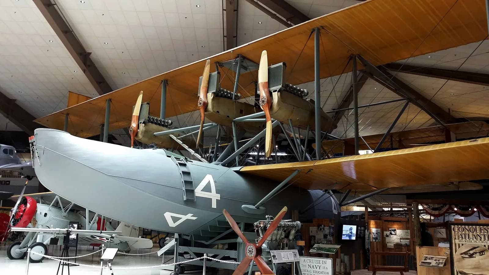 A huge grey flying-boat hull with a large white number 4 sits under long varnished wooden wings in a museum hangar, with three engines and wooden propellers mounted between the wings. A spare engine and propeller stand on the floor in front.
