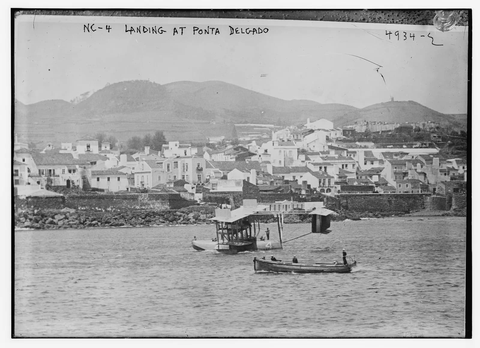 An old glass-plate photograph of a biplane flying boat on calm water with a small launch carrying sailors in front of it. Behind it are a stone seawall, a town of white houses and green volcanic hills. Handwriting at the top reads “NC-4 landing at Ponta Delgado”.