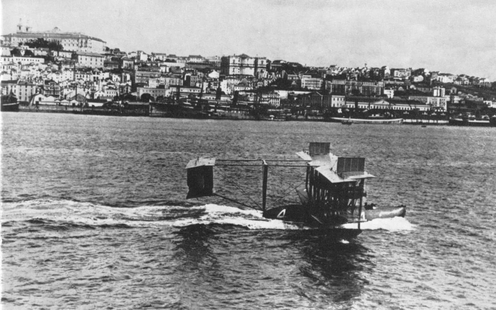 A large biplane flying boat with a boxy tail and the number 4 on its hull speeds across choppy water, throwing up spray. Behind it, across the river, a hillside city of white buildings rises above a waterfront lined with ships.