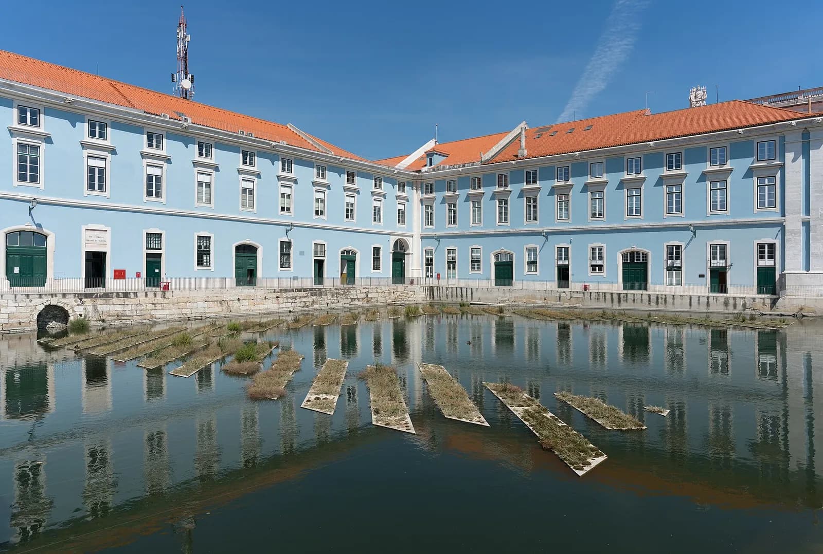 Pale blue three-storey buildings with orange tiled roofs around a flooded stone dock, with rows of old timbers lying in the still water.