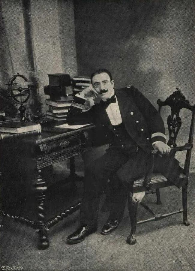 Black-and-white studio photograph of a man with glasses and a moustache in a naval uniform, sitting beside a desk piled with books, with a ringed brass model of the heavens on the desk.
