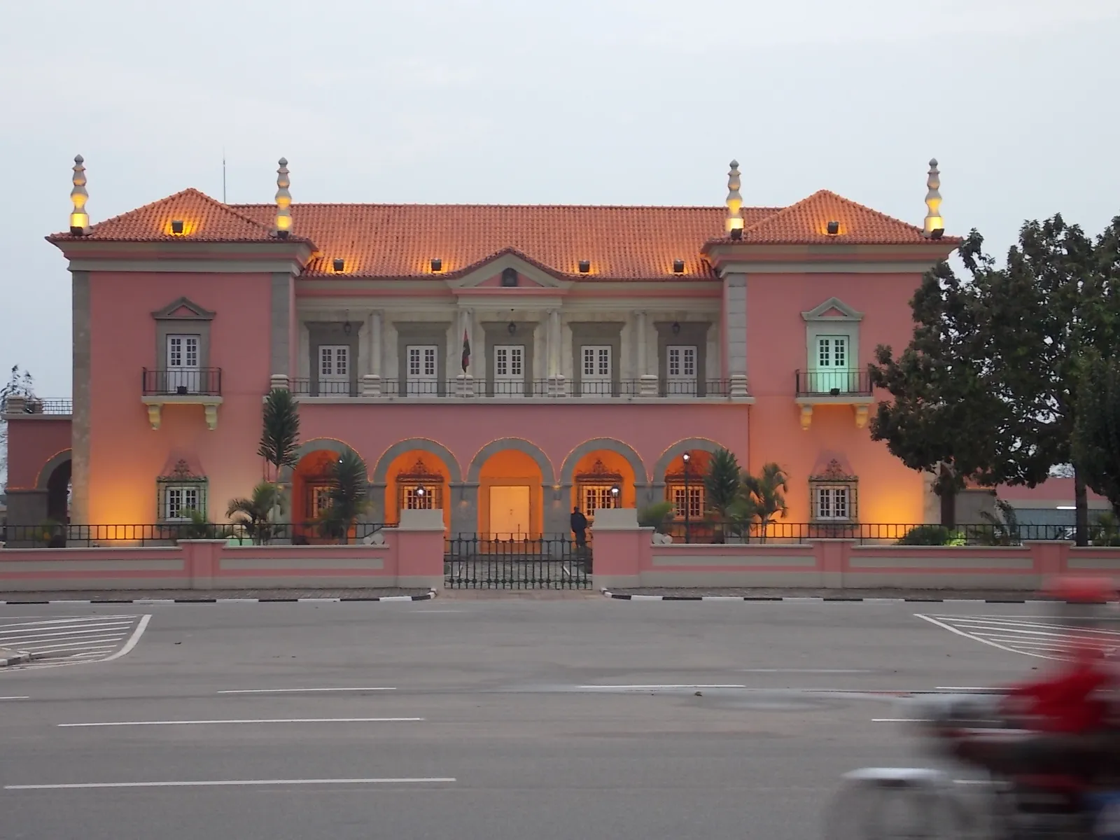 A two-storey pink palace with grey stone trim, an orange tiled roof topped by lit stone finials, and an arcaded ground floor behind a low wall, seen across a wide paved road at dusk.