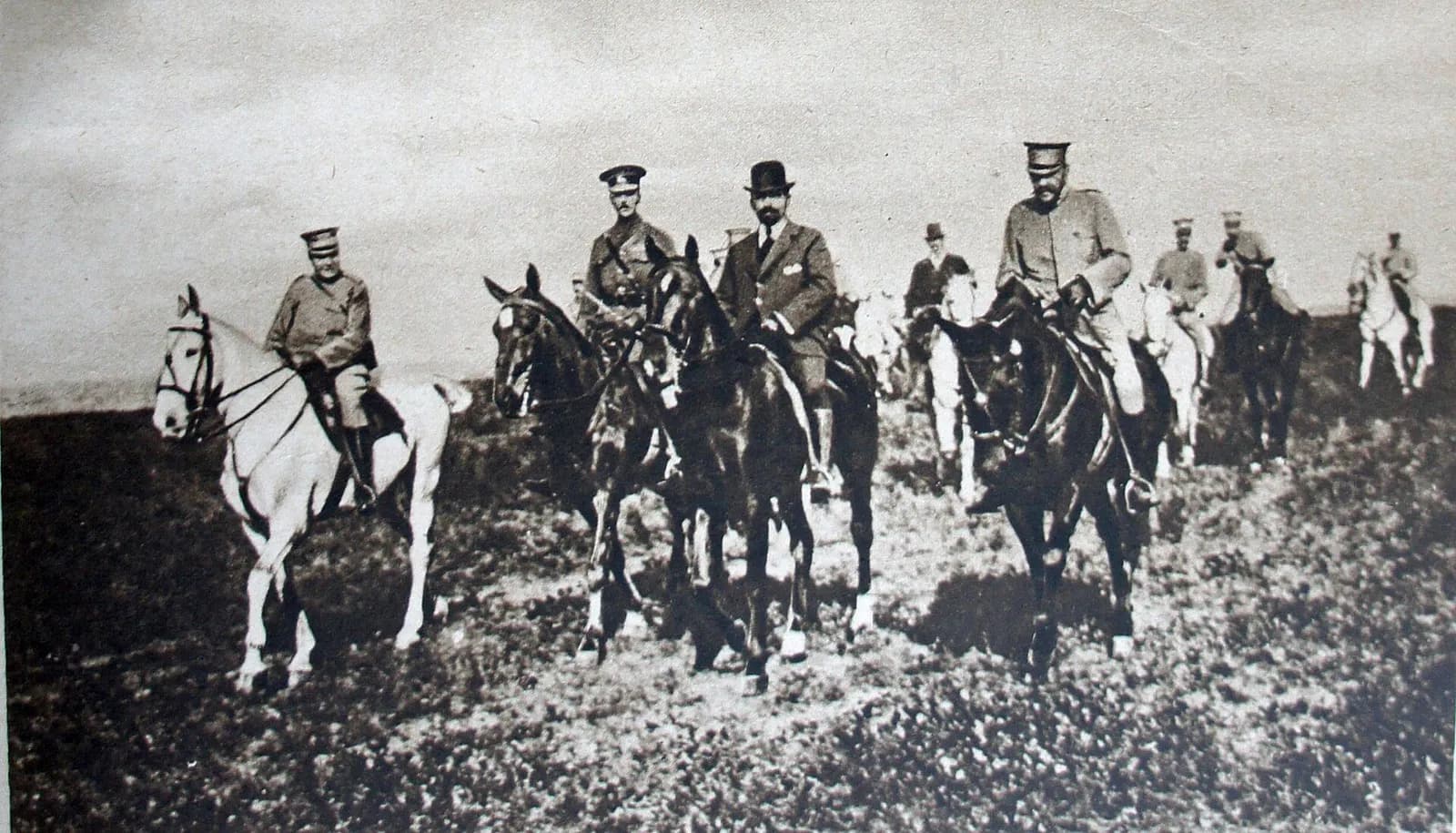 A group of riders on horseback crosses an open field towards the camera. Most wear military uniforms and peaked caps; one, in the centre, wears a dark civilian suit and bowler hat.
