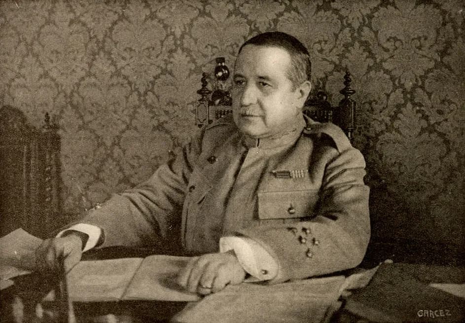 A heavy-set man with short hair and a moustache, in a buttoned military tunic with a row of medal ribbons, sits at a desk covered with open papers, both hands resting on them. Behind him is patterned damask wallpaper and the carved back of a tall chair.