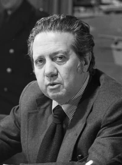 Black-and-white head-and-shoulders photograph of a heavy-set man with swept-back hair, in a textured jacket, striped shirt and dark knitted tie, speaking as he looks slightly off camera.