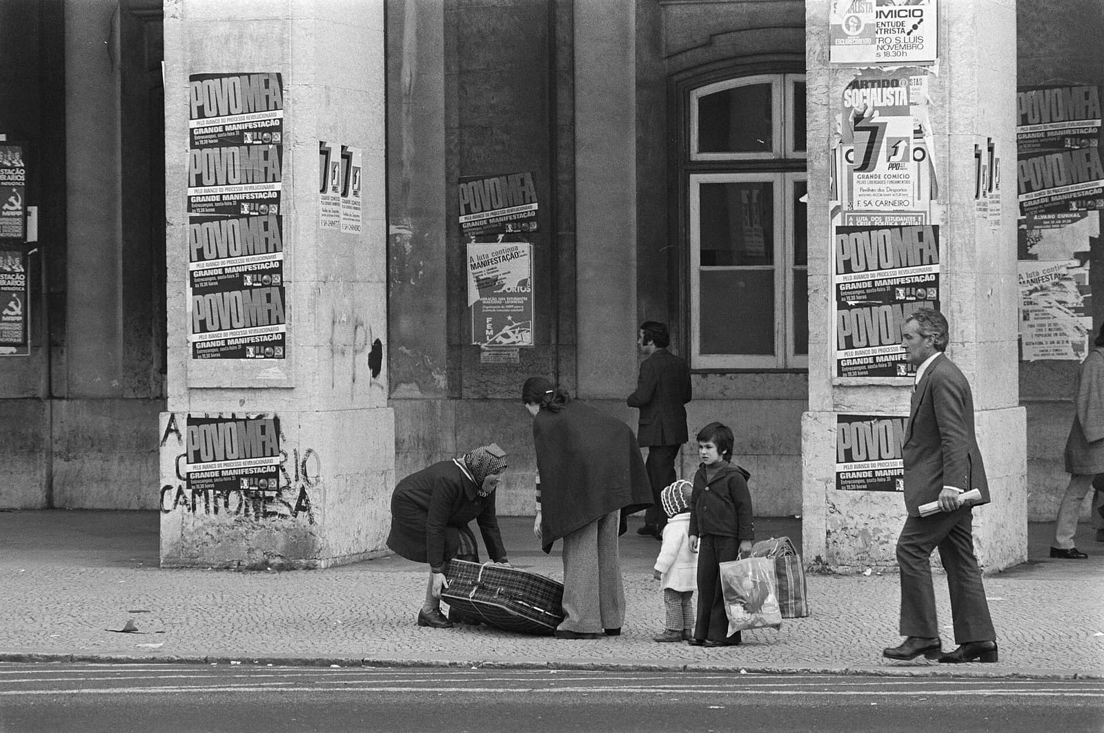 Stone pillars of a building plastered with political posters, many of them printed in large letters with the words Povo MFA, and a painted slogan below. On the pavement an older woman in a headscarf and a younger woman bend over a large checked suitcase beside a small child and a boy holding bags, while a man in a suit walks past.