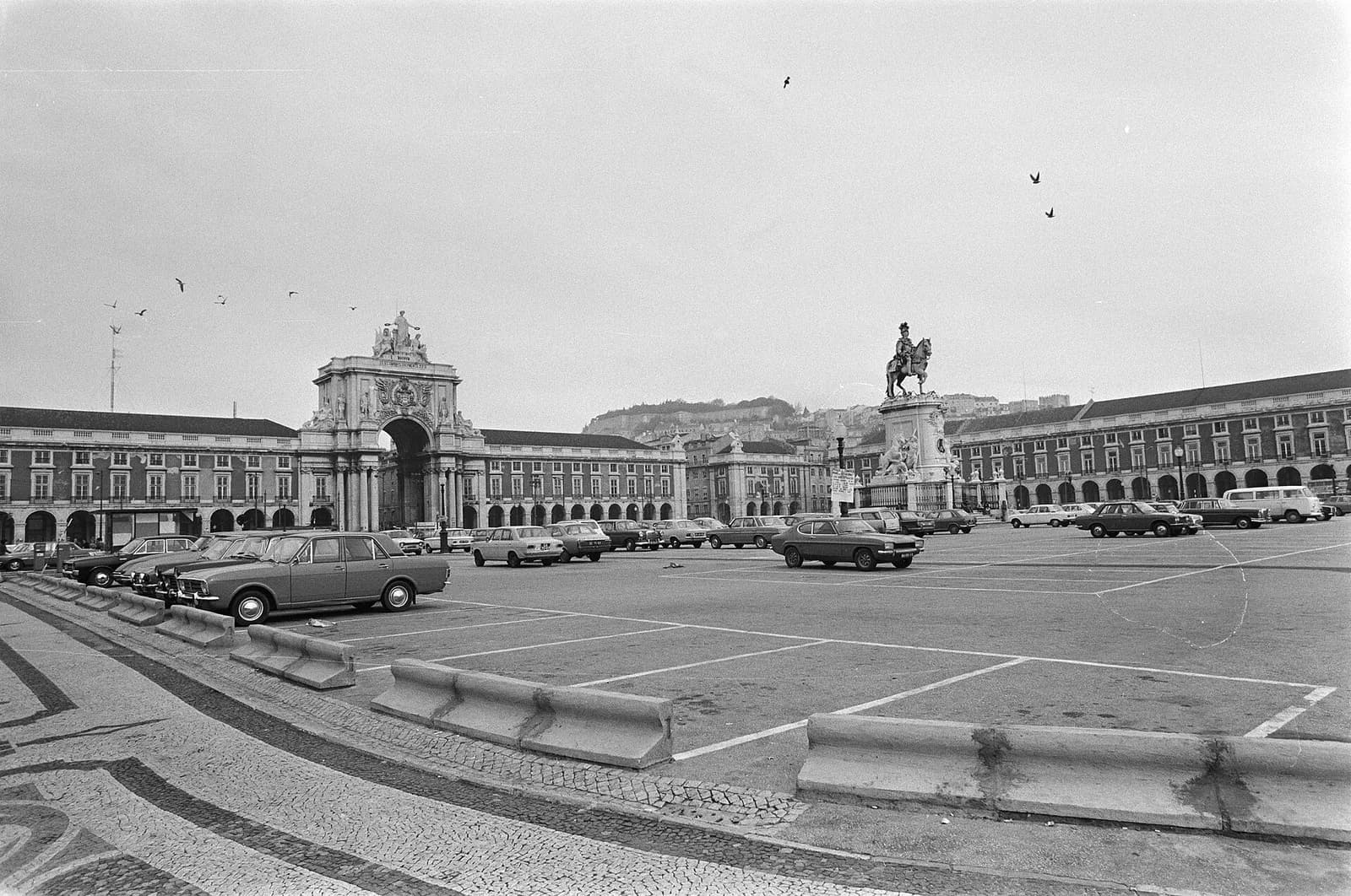 A black-and-white view across a wide paved square used as a car park, with rows of 1970s cars, low concrete barriers in the foreground and long arcaded buildings on three sides. A triumphal arch rises at the far end and an equestrian statue stands to the right.