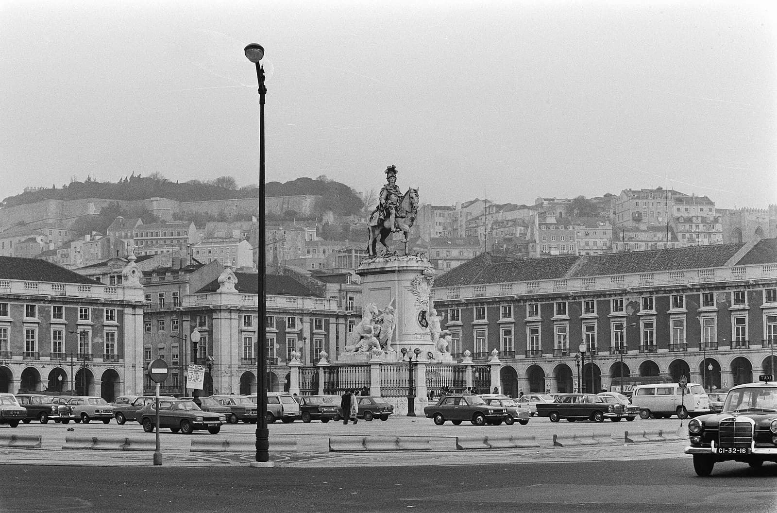 Across a wide square lined with parked cars, an equestrian statue on a tall plinth stands in front of long arcaded buildings, with the hillside of old Lisbon rising behind to the walls of a castle.