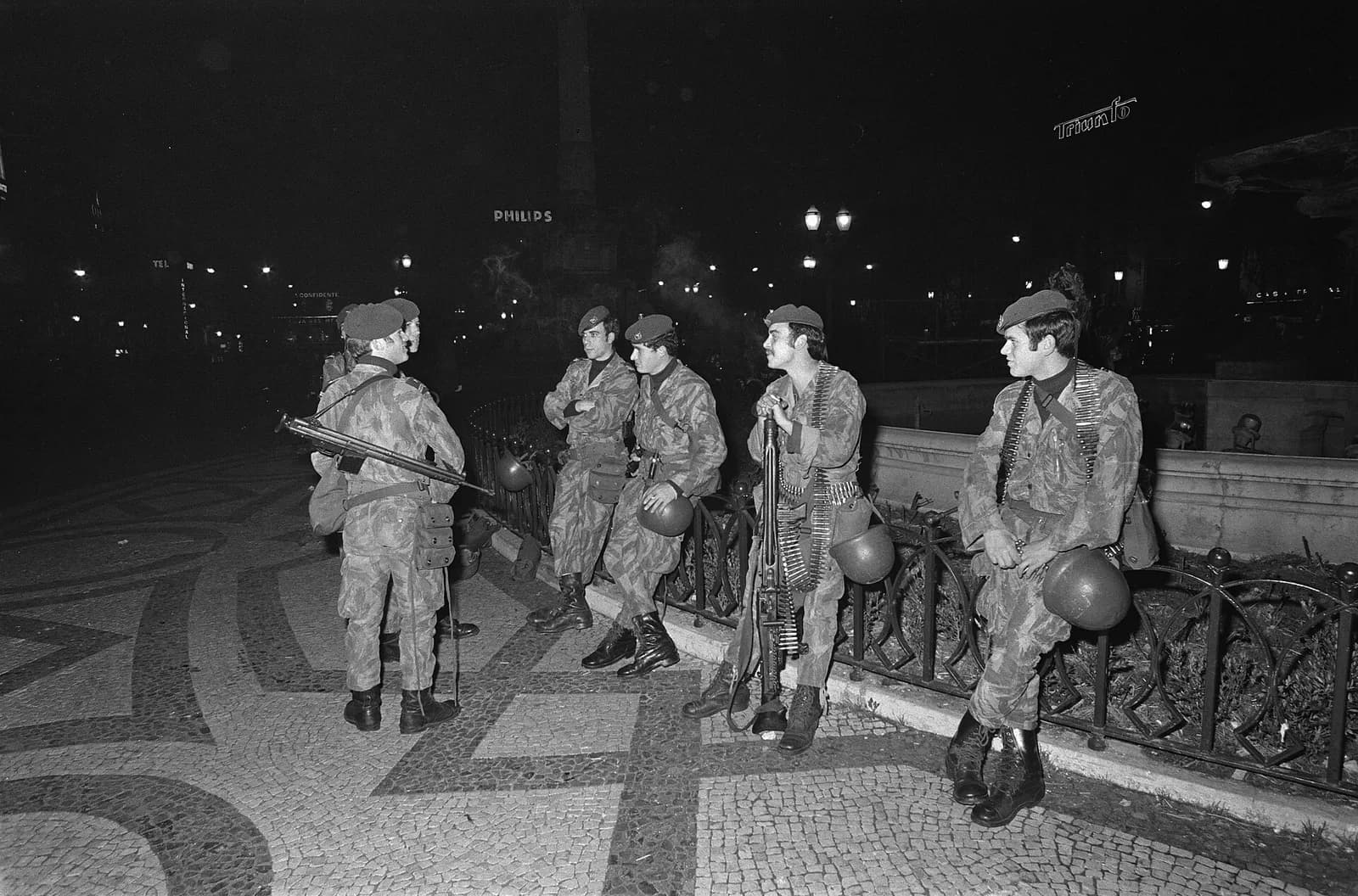 A group of soldiers in camouflage uniforms and berets, with rifles, a machine gun and ammunition belts, stand and lean on a low railing beside a patterned cobbled pavement in a city square at night, some holding their helmets, with street lamps and illuminated signs behind them.