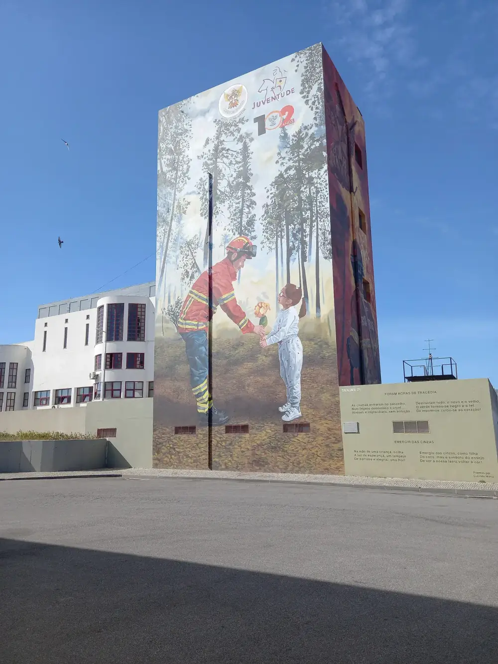 A tall tower painted with a mural of a firefighter in protective gear handing a small flower to a young girl among tall pines, next to a low wall carrying two short poems, under a clear blue sky.
