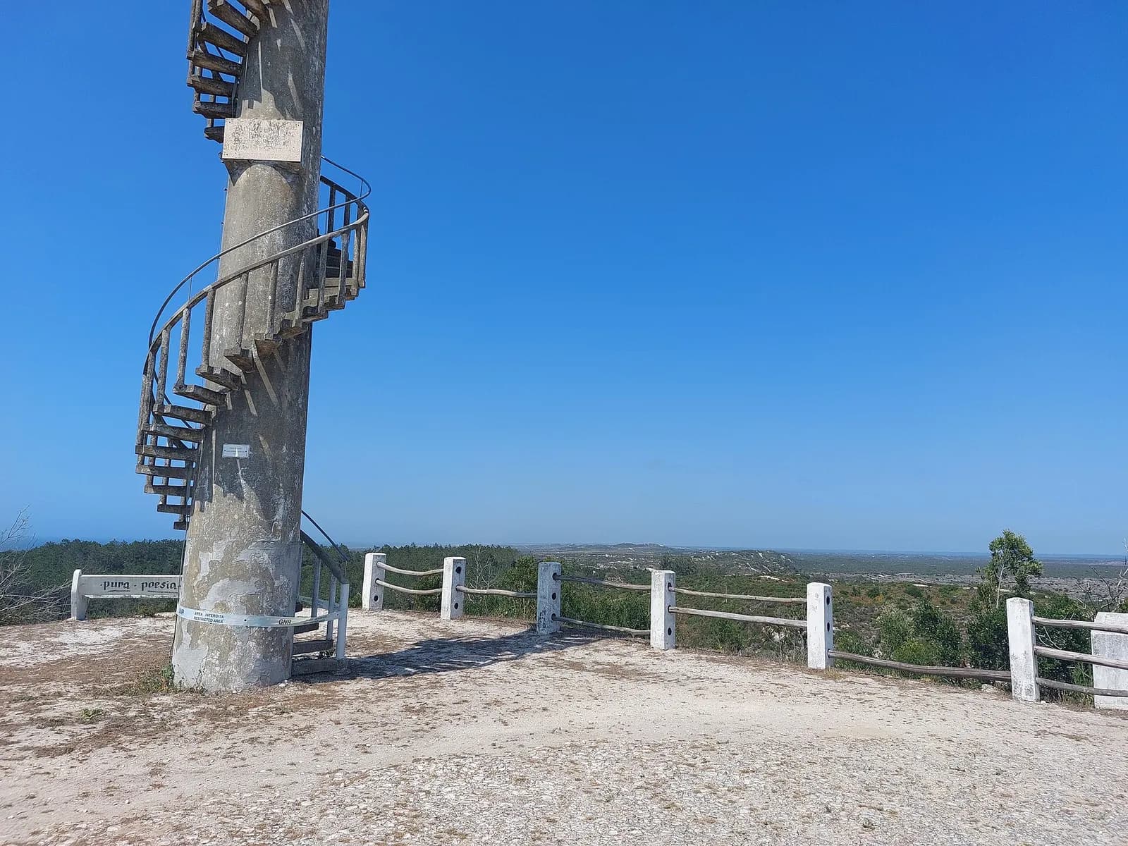 A concrete fire lookout post with a spiral staircase on a sandy hilltop, behind a low wooden fence, overlooking low green scrub and young trees that stretch to a flat horizon under a clear blue sky.