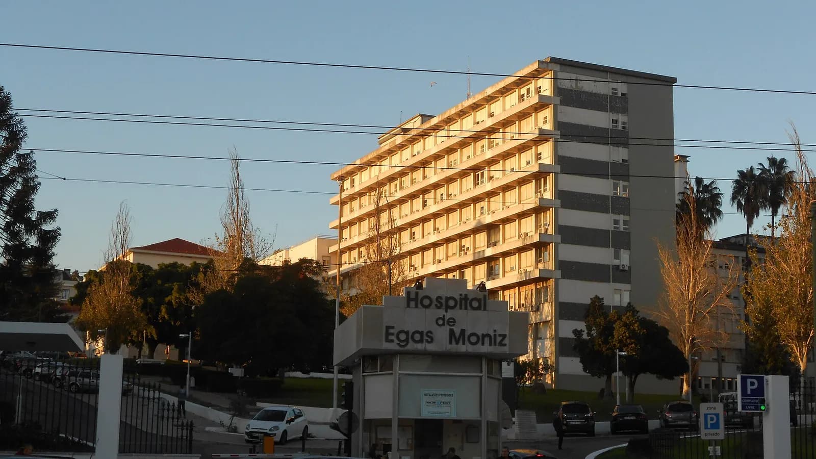 A tall modern hospital block lit by low evening sun behind trees, with a small gatehouse in front bearing the words Hospital de Egas Moniz.
