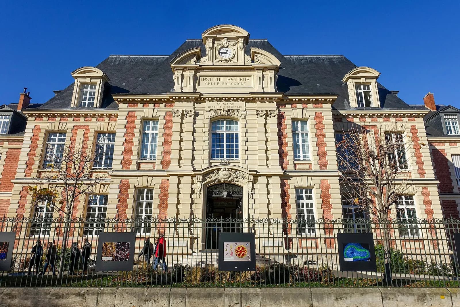 A symmetrical brick and stone building with a slate roof and a clock in its central pediment, inscribed Institut Pasteur, Chimie Biologique, behind iron railings with posters on the fence.