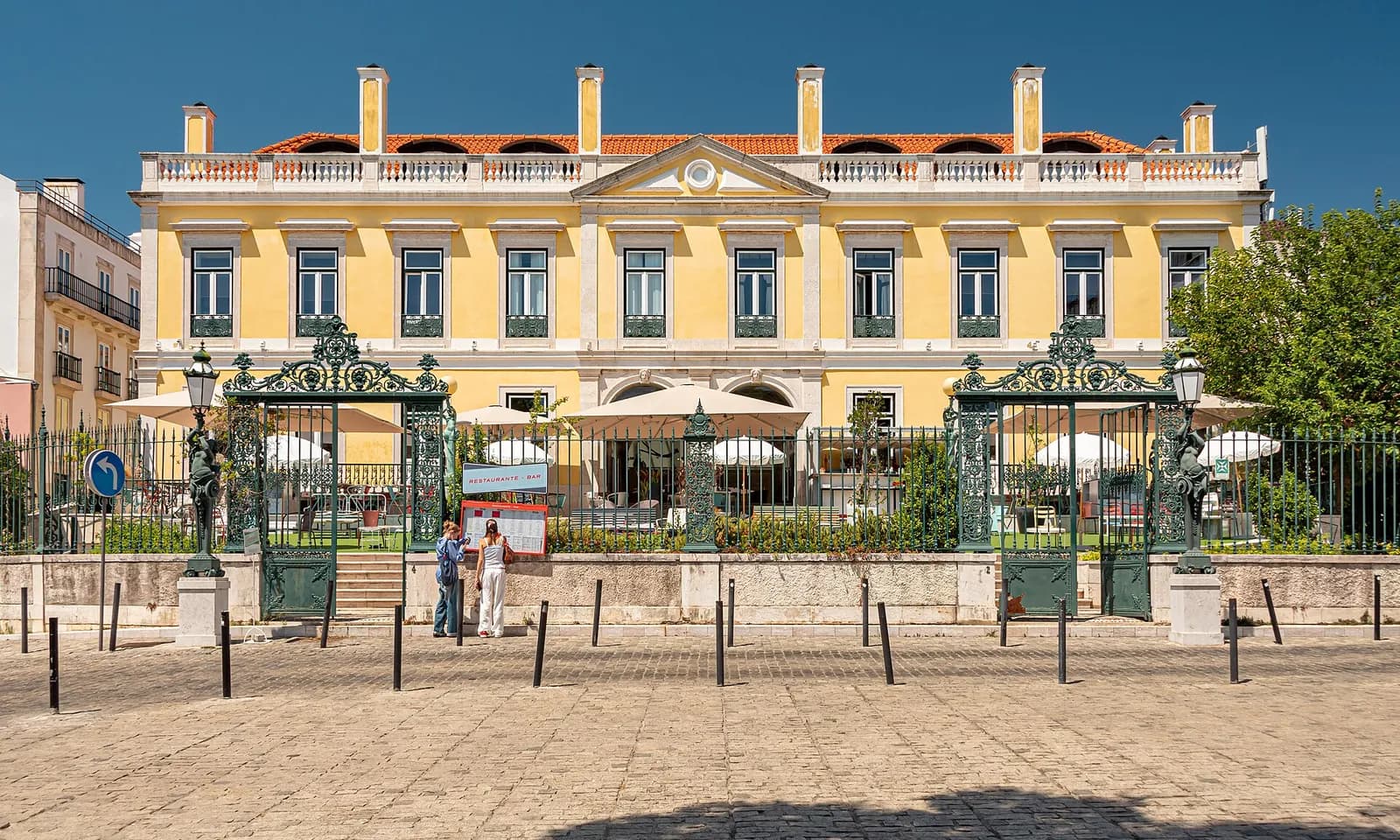 A yellow two-storey palace with white trim and a pediment, seen across a cobbled square through ornate green iron gates, with café umbrellas in its front garden.
