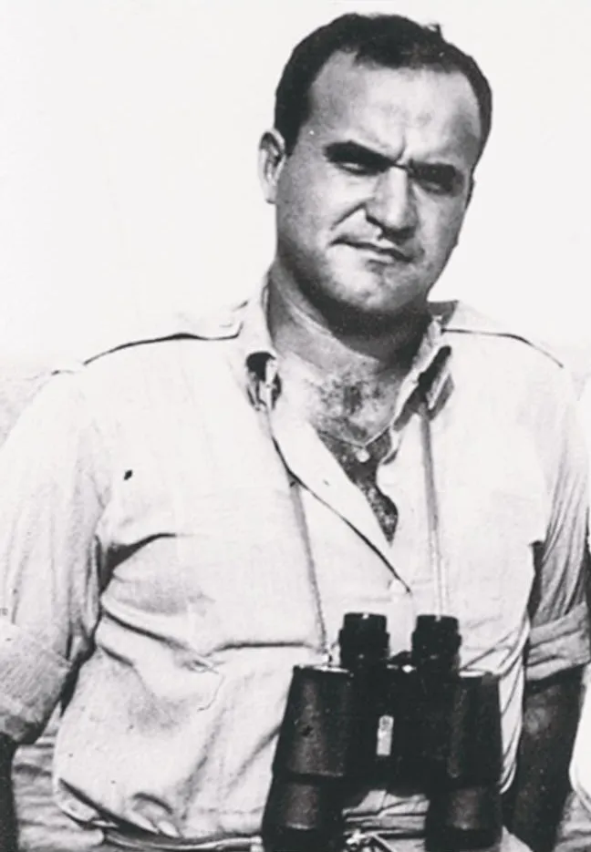 Black-and-white photograph of a stocky, dark-haired man in a light open-necked shirt with rolled sleeves, a pair of binoculars hanging on his chest, frowning slightly at the camera.