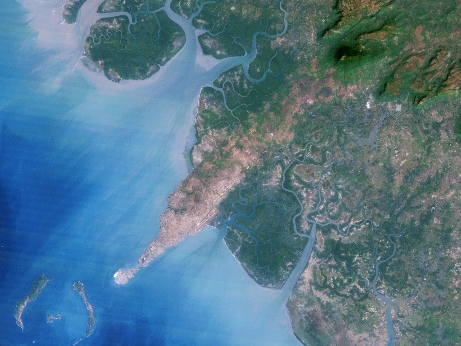 Satellite view of a coast: a long, narrow built-up peninsula reaches south-west into blue sea, with a pale runway halfway along it, mangrove swamps and river channels inland, and two long, thin islands and a smaller one offshore.