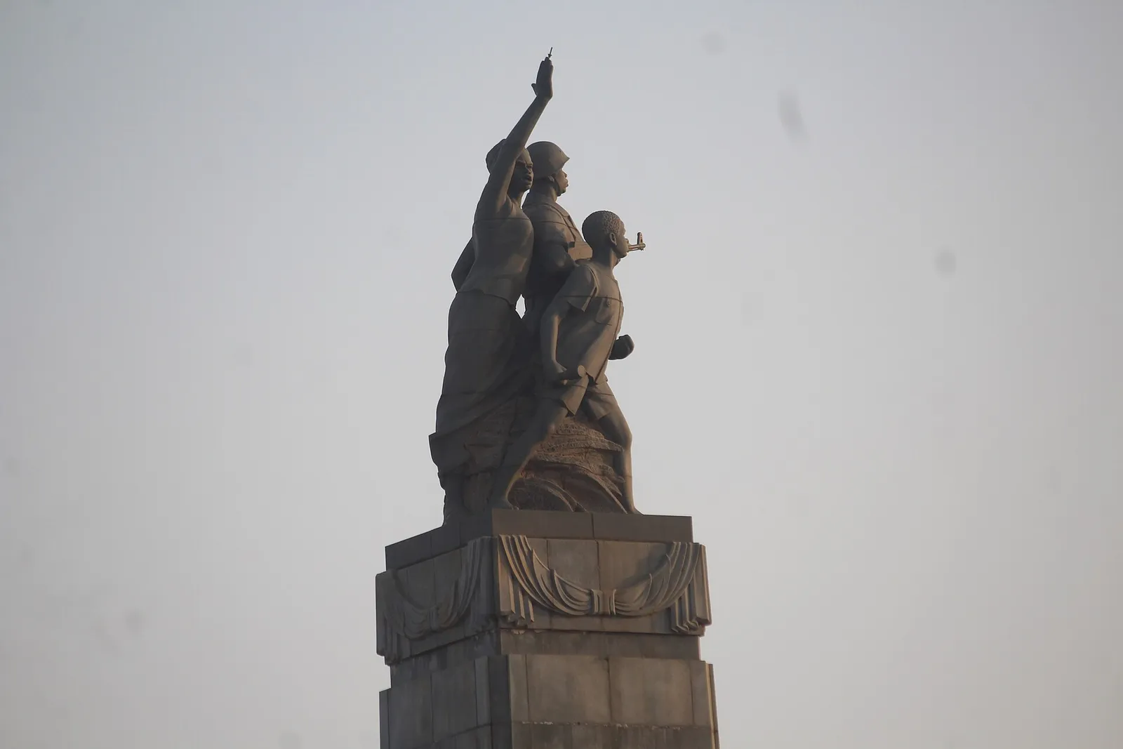 A dark stone statue on a tall plinth against a hazy sky: a woman with her arm raised high, a soldier in a helmet holding a rifle, and a boy striding forward with his fists clenched.
