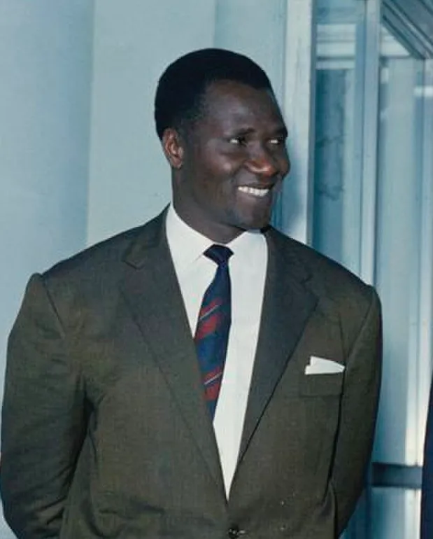 Colour photograph of a smiling man with short hair in a dark olive suit, white shirt and striped tie, a white handkerchief in his breast pocket, looking to one side.