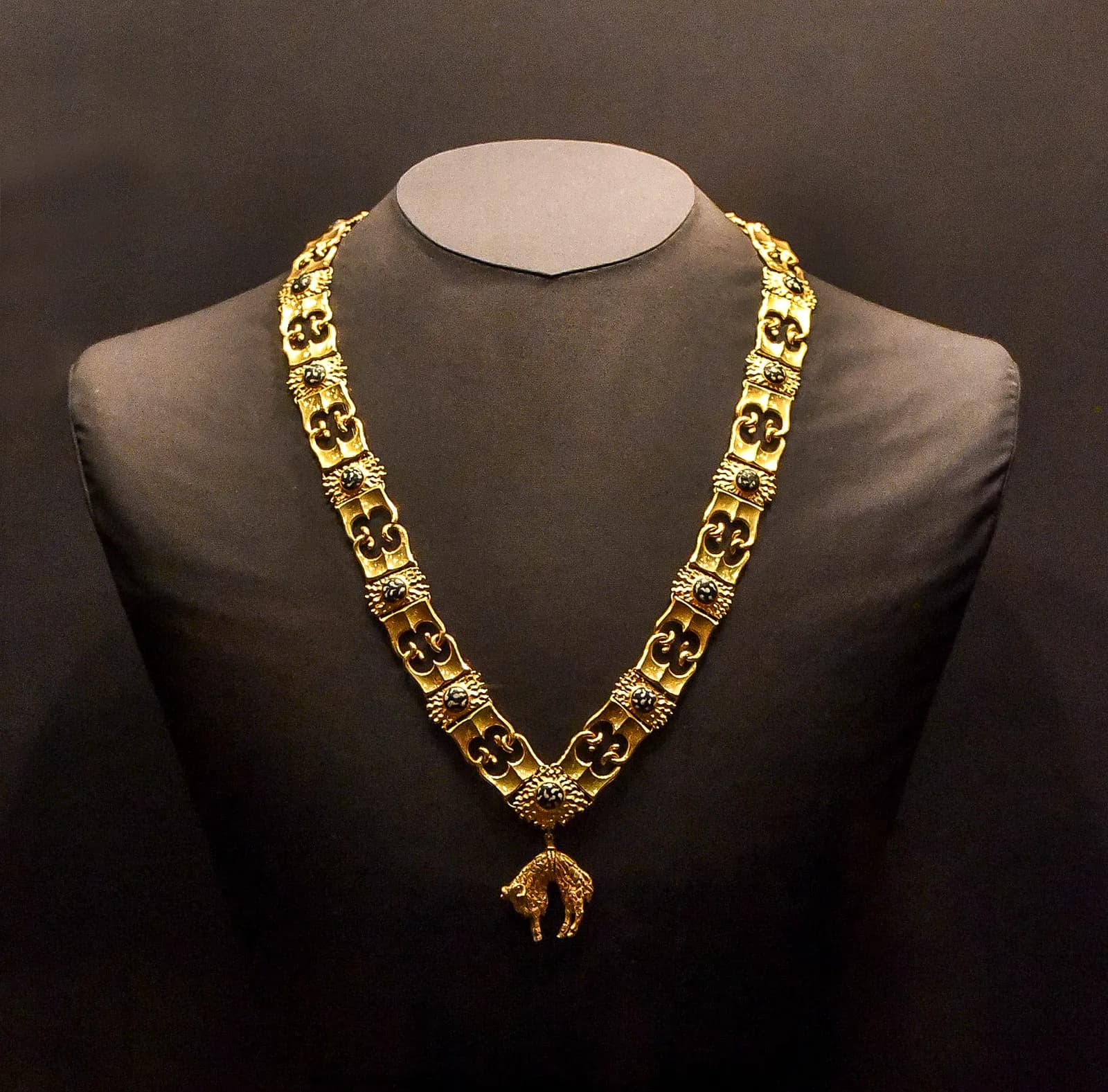 A heavy gold collar displayed on a dark fabric bust. Its links alternate between pairs of curved B-shaped fire-steels and square plaques set with black enamel flecked with white, and a small gold ram hangs from the point at the front.