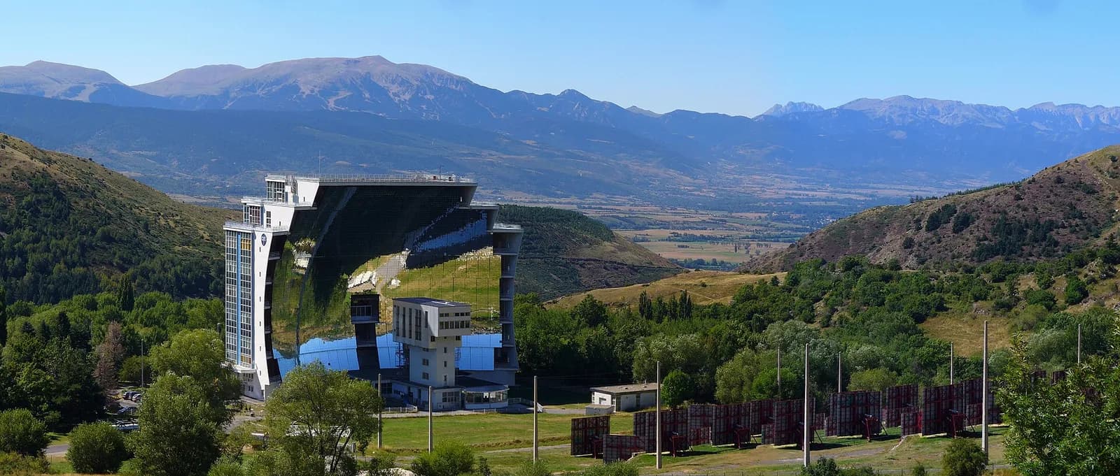 A tall white building whose whole face is a giant curved mirror reflecting the green hills, set in a mountain valley, with rows of flat mirror panels on the slope in front of it.