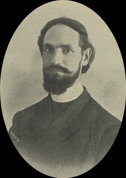 Oval portrait of a man in his late thirties with dark swept-back hair, a full dark beard and moustache, wearing a dark coat over a clerical collar.