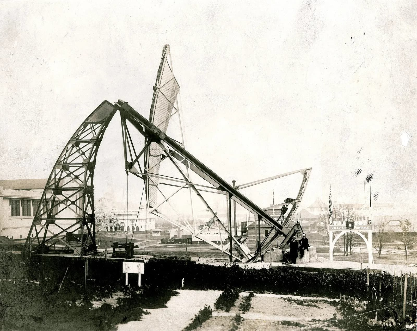 A huge tilted steel frame on open ground: a curved lattice arch supports a long beam, at one end of which rises a tall, narrow reflector panelled with small squares. Two men in dark coats, one standing and one seated, are at its base, and a white arch with flags stands to the right.