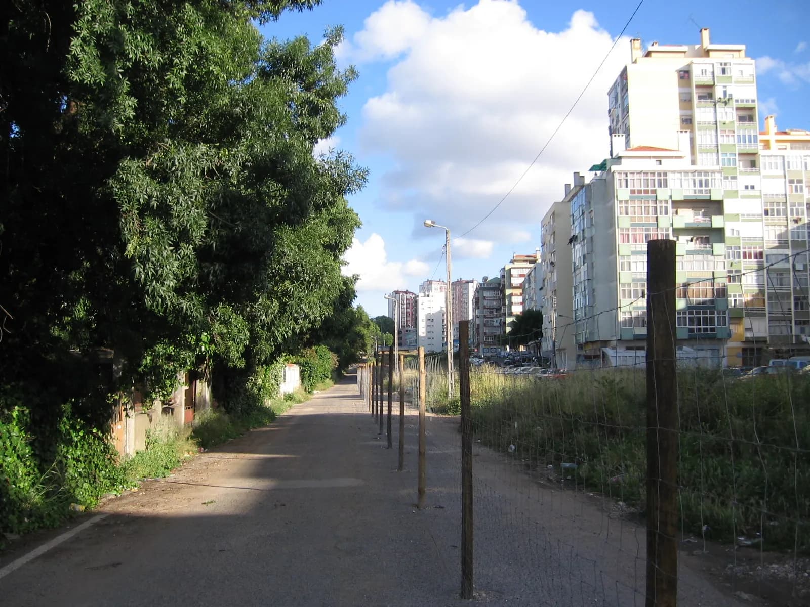 A narrow, partly unpaved road runs between overgrown trees and low walls on the left and a wire fence on wooden posts on the right, beyond which rough ground leads to tall apartment blocks under a blue sky.