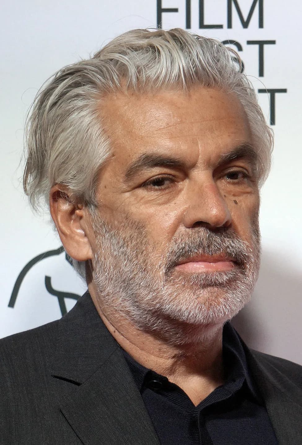 Head-and-shoulders portrait of a man in his sixties with swept-back grey hair and a short grey beard, wearing a dark jacket over a dark shirt, in front of a white festival backdrop.