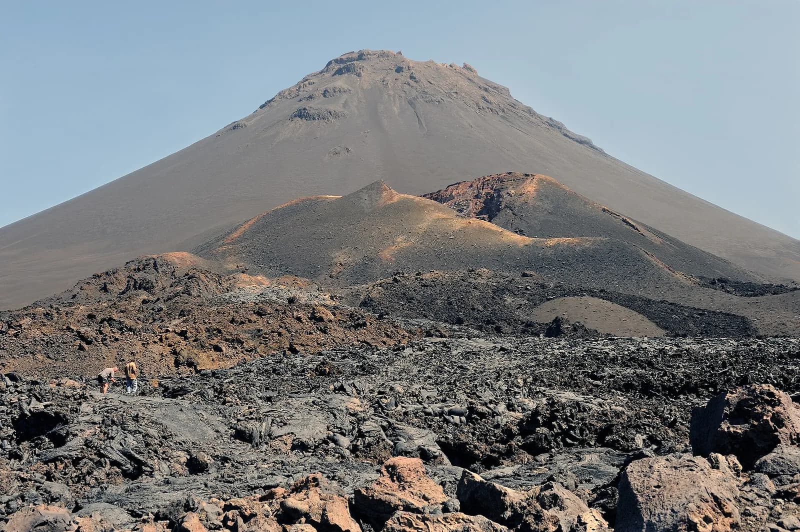 A steep, bare grey volcanic cone rises against a pale sky behind smaller reddish-brown cones and a wide field of black, ropy lava, where two small figures stand on the left.