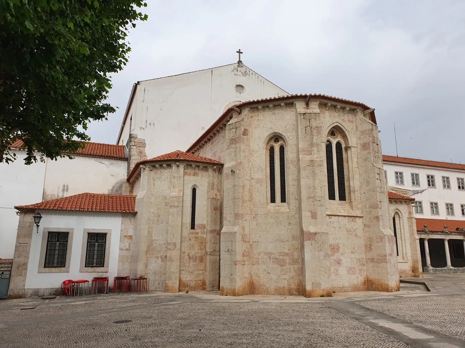 The east end of a medieval stone church with tall narrow Gothic windows and red-tiled roofs, seen across a cobbled square. White later buildings with rows of windows stand on either side.