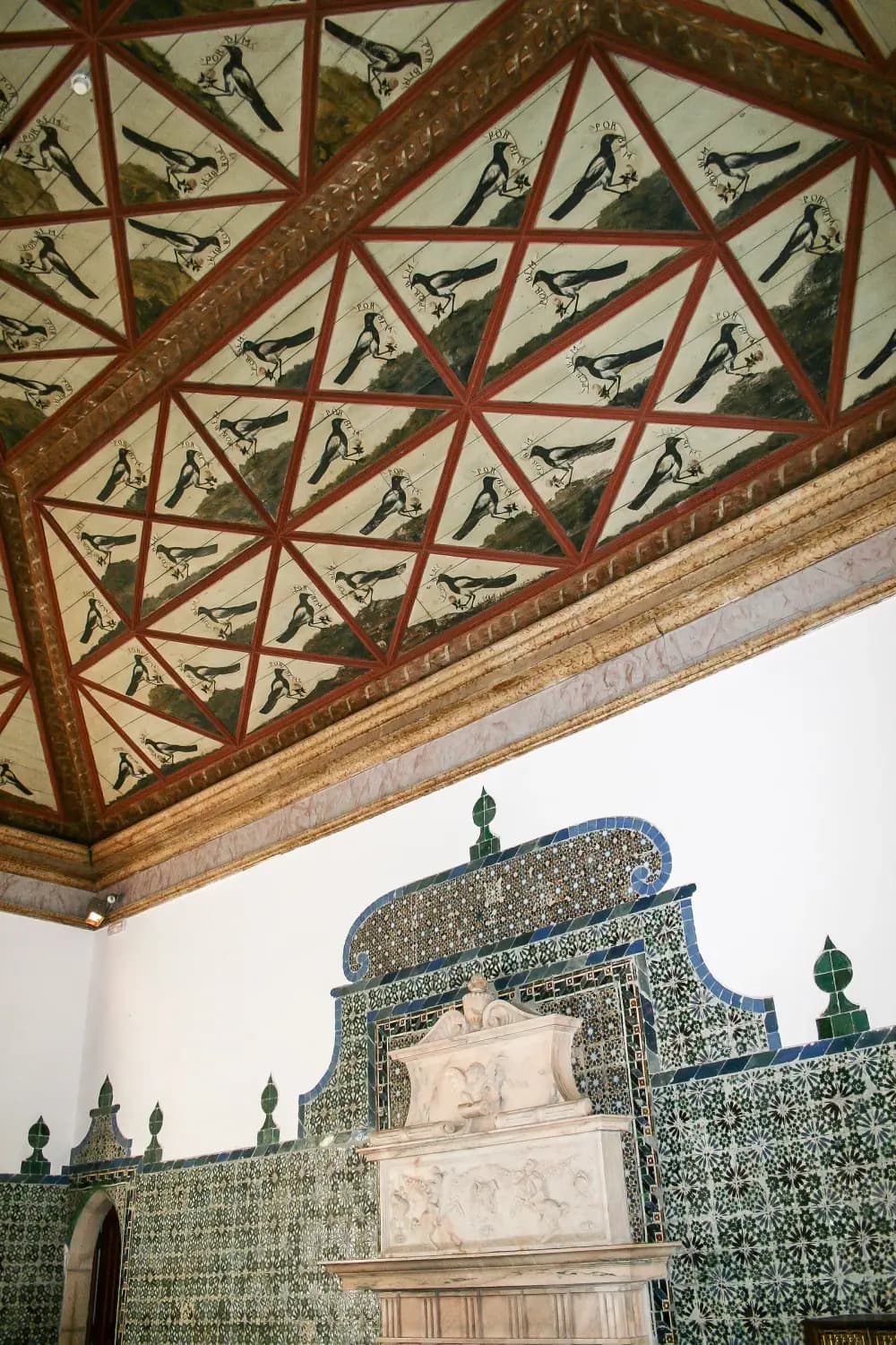 A painted wooden ceiling divided by red ribs into many triangular panels, each showing a black and white magpie holding a small scroll and a flower. Below, a white wall is lined with green and white tiles around a carved white marble fireplace.