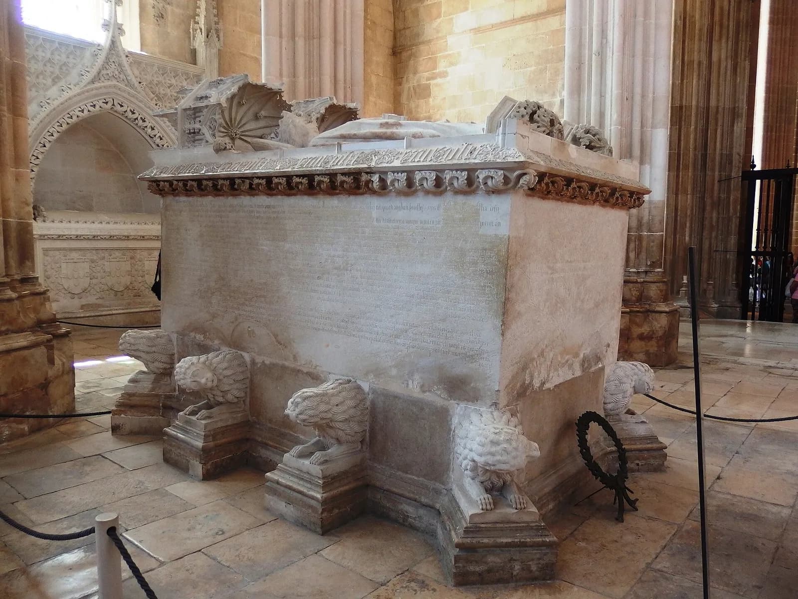 A large pale limestone tomb chest resting on carved lions stands in a Gothic chapel. Carved stone canopies and the tops of two recumbent effigies show above the rim of the lid, and the long side is covered in a carved inscription.