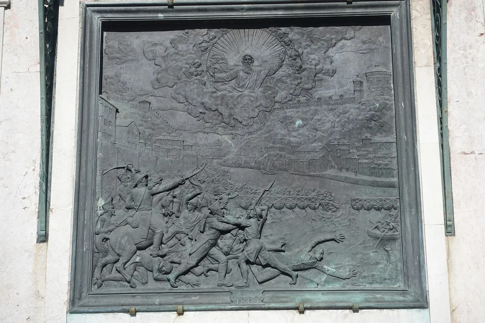 A weathered bronze relief in a metal frame on a pale stone wall. Soldiers on horseback with raised sabres drive a crowd of men towards a river, where people fall from a low bridge and struggle in the water and a boat picks up survivors. A walled town lines the far bank, and a bearded God the Father surrounded by cherubs looks down from the clouds. A small plaque at the bottom reads 29-3 1809.