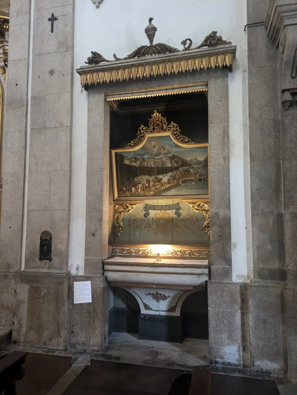 A granite niche in a church wall holds a small altar. Above it, in a gilded frame, a painting shows French cavalry on a quay below a walled town, people struggling on a wooden bridge and in the river, and a holy figure in the clouds. A painted ribbon below reads 29 de Março de 1809.