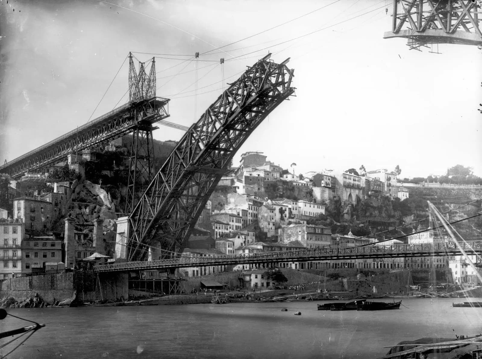 Black-and-white photograph of a half-built iron arch rising steeply from one bank of a river, its unfinished end held by cables from a tall lattice tower on the completed approach deck. Below it a slender suspension bridge with a lattice railing crosses the river in front of houses on a steep hillside.