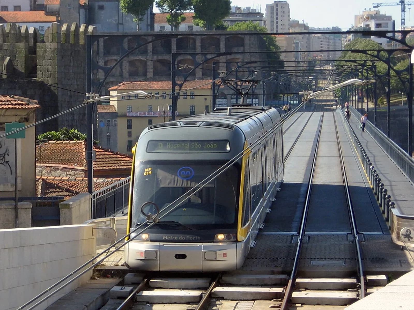 A grey and yellow light-rail train marked Metro do Porto, showing the destination D Hospital São João, comes off a bridge deck with two tracks under overhead wires hung from decorative iron arches. Pedestrians walk on a railed footway to the right, and an old crenellated stone wall rises on the left.