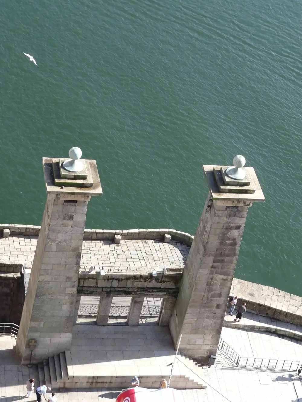 Seen from high above, two tall granite pillars topped with stone balls stand at the edge of a paved quay beside green water, with a low stone façade with three openings between them and a few people walking below.