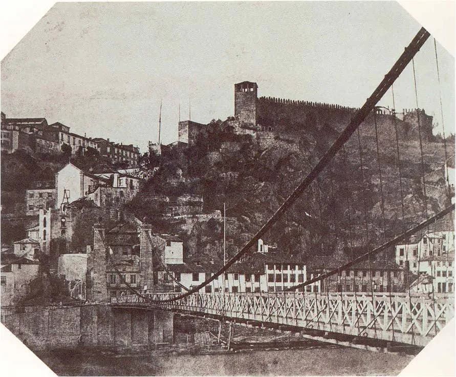 An early sepia photograph, cut to an octagon, of a suspension bridge with a lattice railing crossing a river. Its cables sweep down from the top right, and behind it houses and a crenellated wall with a tower climb a rocky hillside.