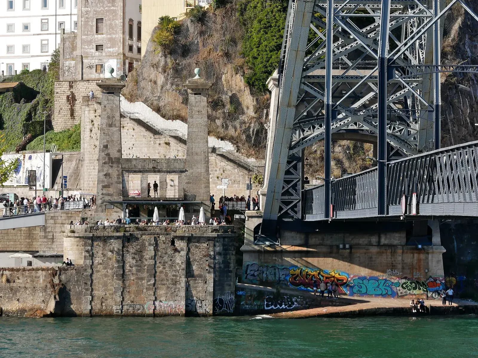 Two tall stone pillars topped with balls rise from a stone pier at the edge of a green river. Between them is a low stone facade with a terrace where people stand under white parasols. To the right, the grey iron frame of a large bridge meets the bank, and graffiti covers the wall below it.