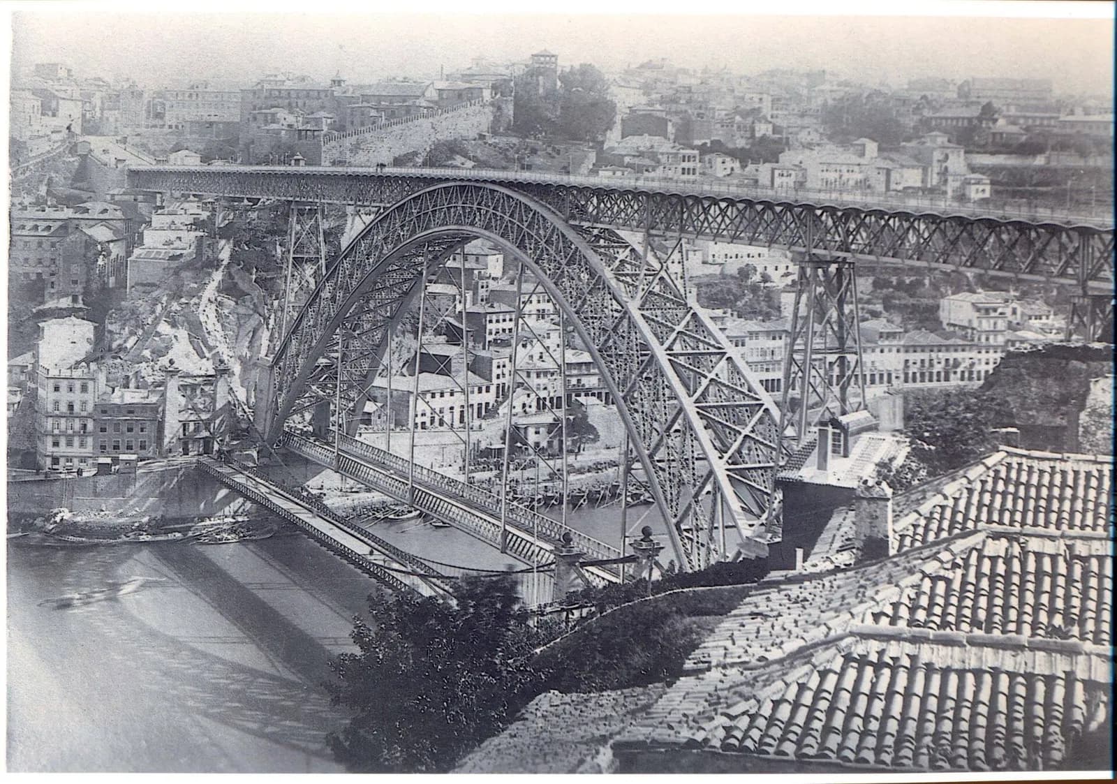 A black-and-white photograph taken over tiled rooftops. A huge iron arch spans a river and carries a high deck across to a hillside city. Low down, slender decks with lattice railings cross the river beneath the arch, and two small stone towers topped with balls stand at their near end.