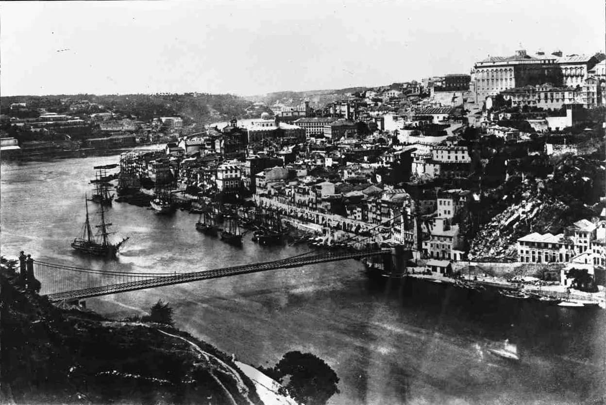 An old black-and-white photograph taken from a hillside above a wide river. A suspension bridge with a lattice railing crosses from two tall stone towers at the lower left to the far bank, where tall houses crowd the slope above a quay lined with sailing ships.