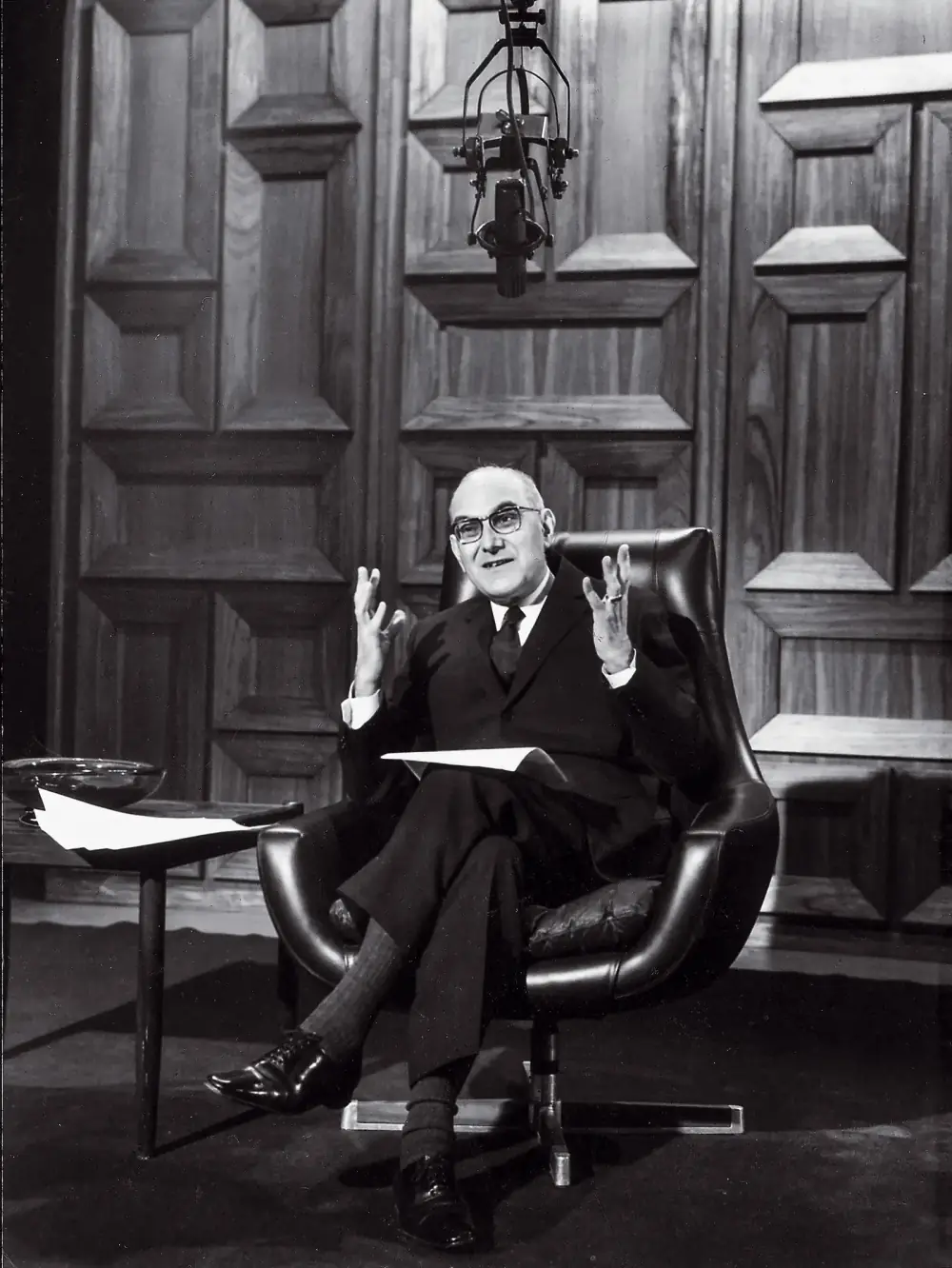 A bald man in glasses and a dark suit sits in a leather swivel chair in a wood-panelled television studio, gesturing with both hands, papers on his lap, a microphone hanging above him.