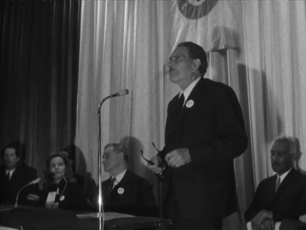 A grey-haired man in a dark suit, holding his glasses and wearing a lapel badge, stands by a microphone on a curtained stage, with a woman and several men seated at a table beside him.