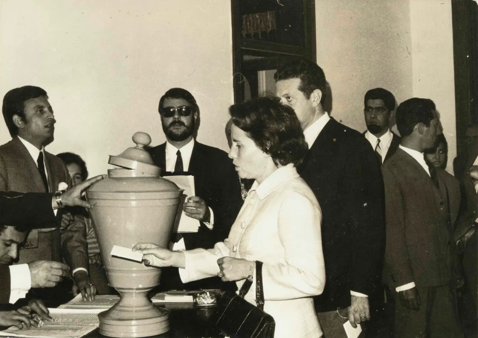 A woman in a pale jacket with a handbag holds a ballot paper at a tall urn-shaped ballot box, a tall dark-haired man in a suit behind her, with officials and onlookers around the table.