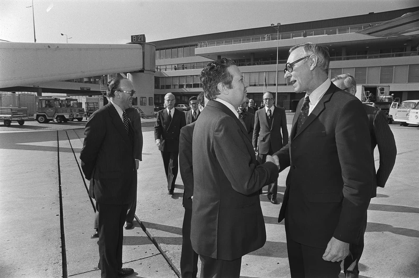 Black-and-white photograph of two men in dark suits shaking hands on an airport apron, with an aircraft boarding bridge marked B21, a terminal building and several officials behind them.