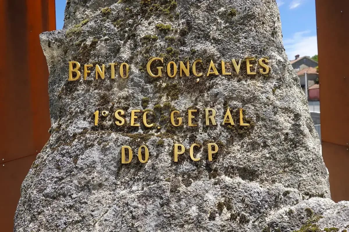 A rough, mossy stone boulder with gilt metal letters reading Bento Gonçalves, 1º Sec. Geral do PCP, framed by two rusted steel panels against a blue sky.