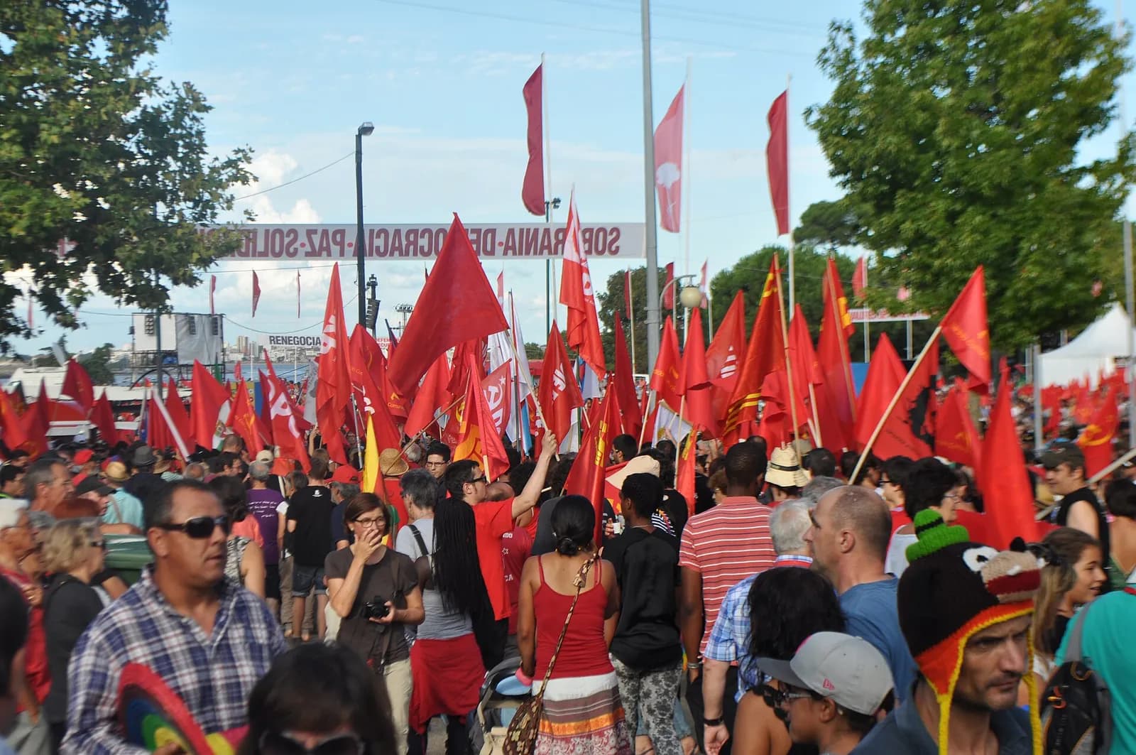 A large crowd in summer clothes walks among dozens of red flags, some with the hammer and sickle and some with the letters PCP, under a banner reading Soberania, Democracia, Paz, Solidariedade.