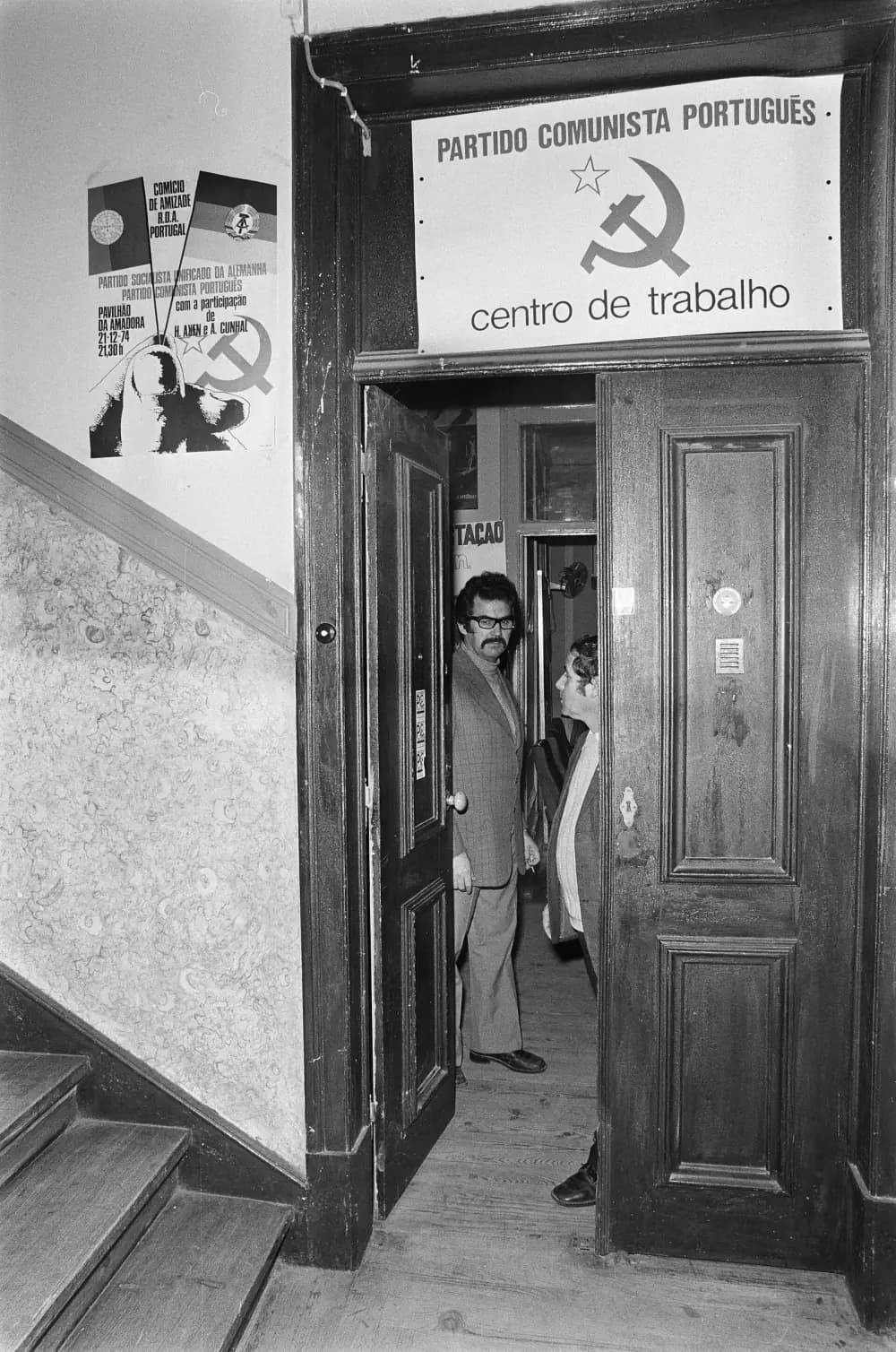Two men stand in the open double doorway of a flat off a staircase. A printed sign above the door reads Partido Comunista Português, centro de trabalho, with a hammer and sickle and a star; a poster on the wall advertises a friendship rally with the East German party and A. Cunhal on 21 December 1974.