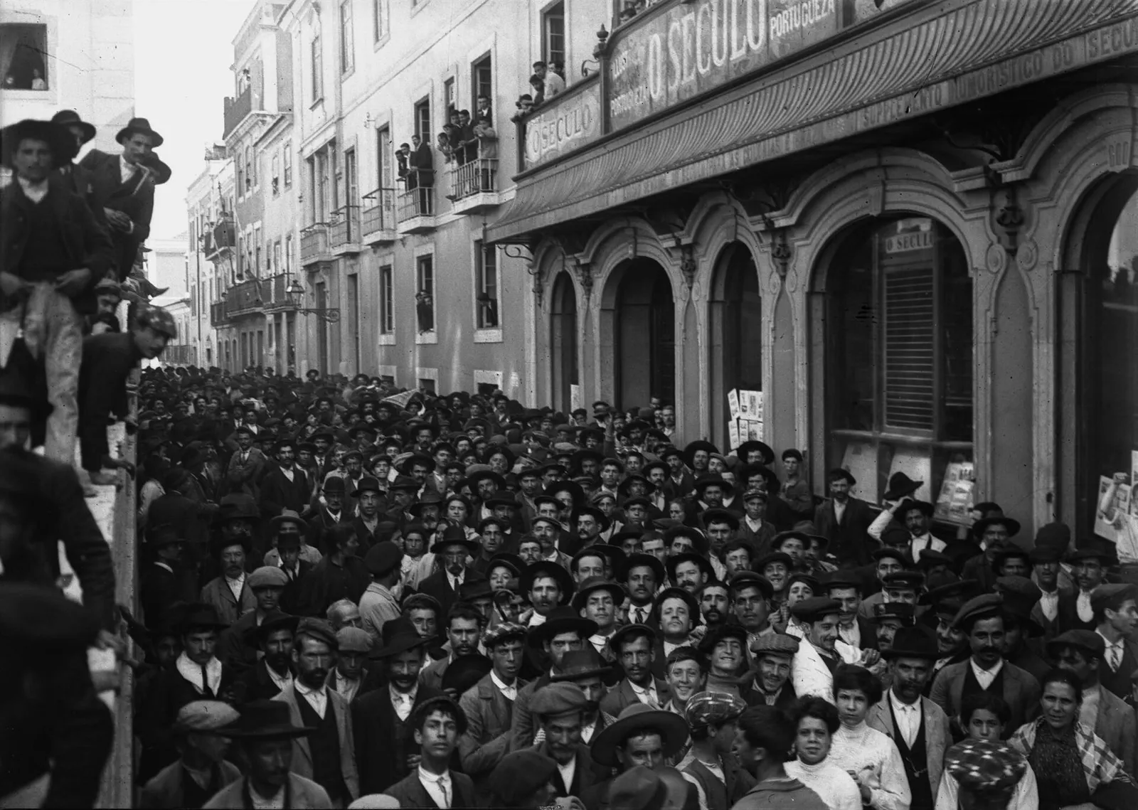 A dense crowd of men in caps and broad-brimmed hats, with a few women, fills a narrow street in front of a newspaper office whose arched shopfront and balcony carry the name O Século. Onlookers lean from balconies and climb a wall on the left.